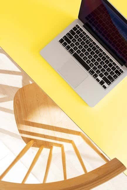 Laptop on yellow desk with wooden chair in bright, modern workspace.